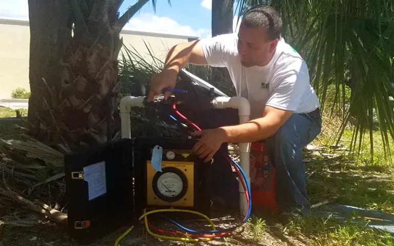 Man in white shirt working on machinery outdoors, near a tree. Yellow gauge visible.
