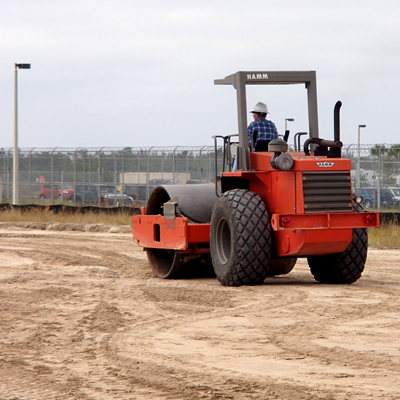 A construction worker operates a red roller machine, compacting soil on a worksite.