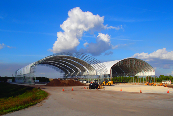 Large white and metal arched structures under a partly cloudy blue sky, with construction equipment and dirt piles.