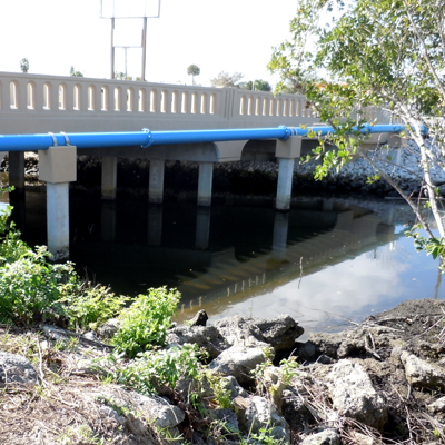 A bridge with blue water pipes, concrete supports, and a dark water canal.