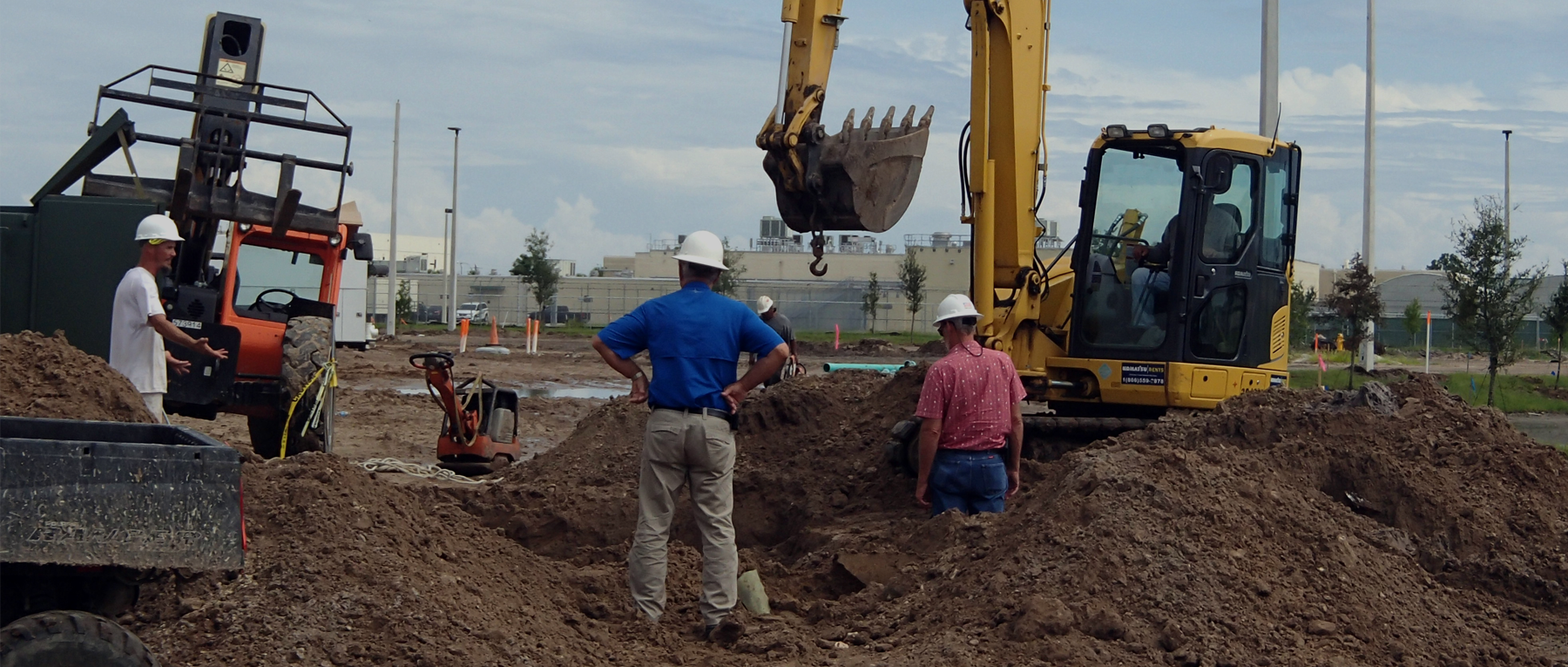 Construction site with yellow excavator, workers in hard hats, and piles of dirt.