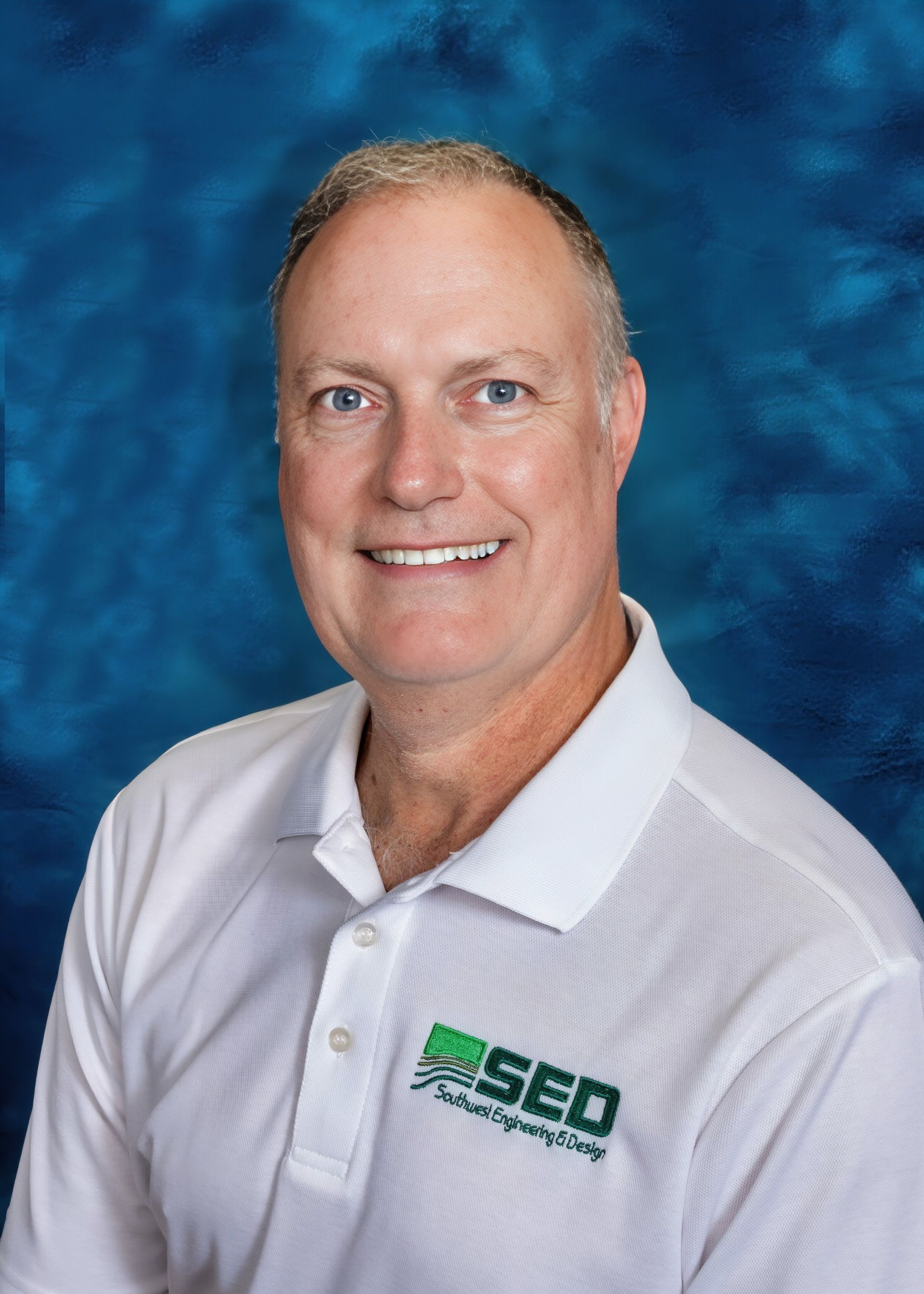 Man wearing white polo shirt smiles. Background is blue. SED logo visible on shirt.