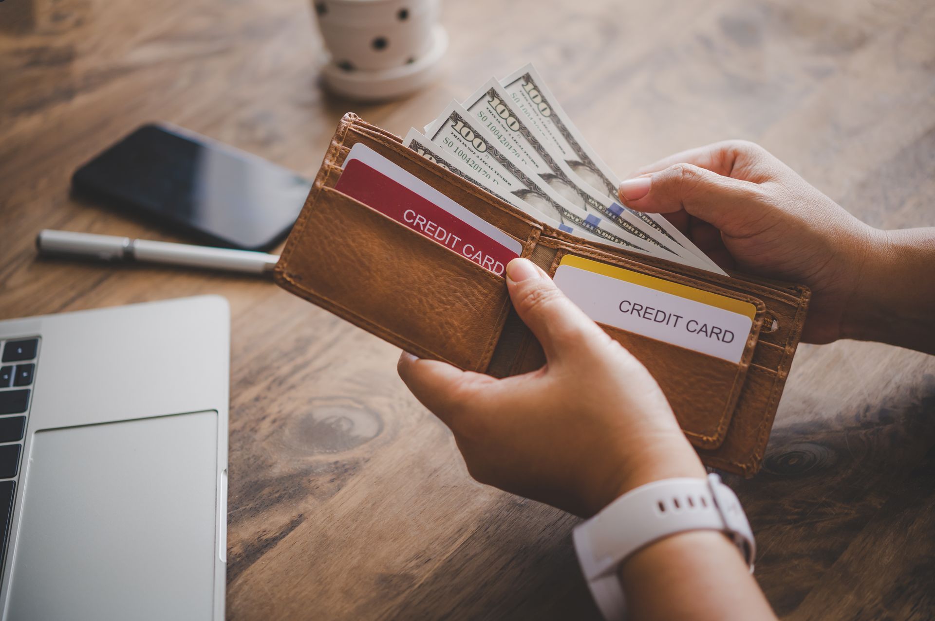 Hands holding a brown leather wallet with cash and credit cards visible on a wooden surface 