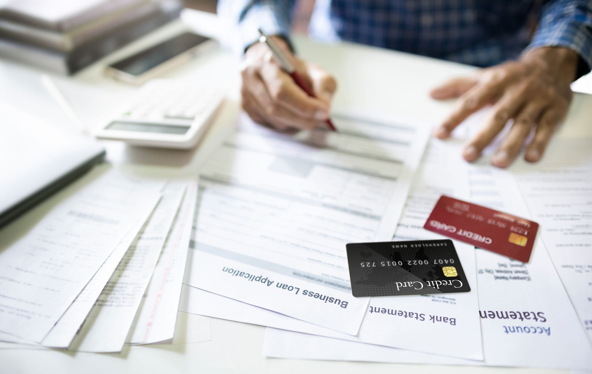 A person writes on financial documents at a desk, with credit cards, a calculator, and papers nearby.