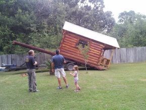 A group of people are standing in front of a log cabin on a trailer.