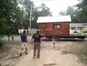 A group of people are standing in front of a log cabin on a trailer.