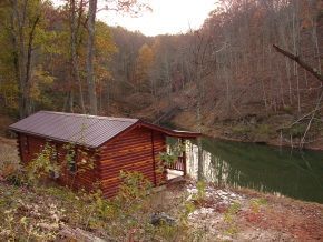 A small log cabin is sitting next to a river in the woods.