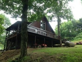 A cabin with a large deck is surrounded by trees and grass.