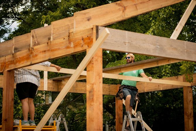 Workers assembling a wooden frame structure outdoors, with two people on ladders under exposed beams.