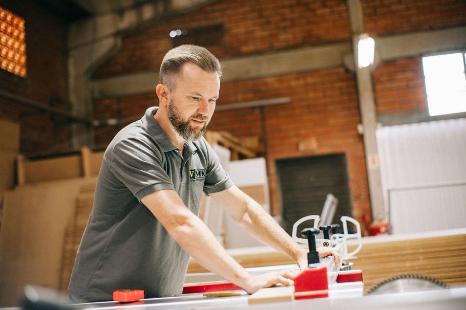 Man in gray shirt measuring wood at a workbench in a brick workshop