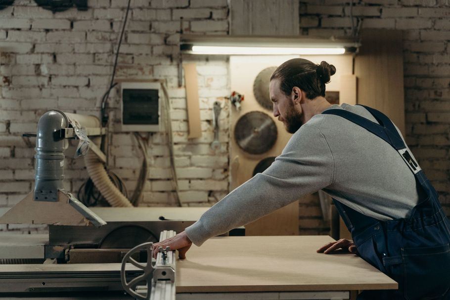 Woodworker in blue overalls sanding wood at a workbench in a brick-walled workshop