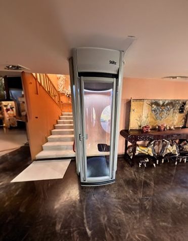 A residential hallway with wooden doors and floors, featuring a modern, cylindrical glass home elevator as a central focus.