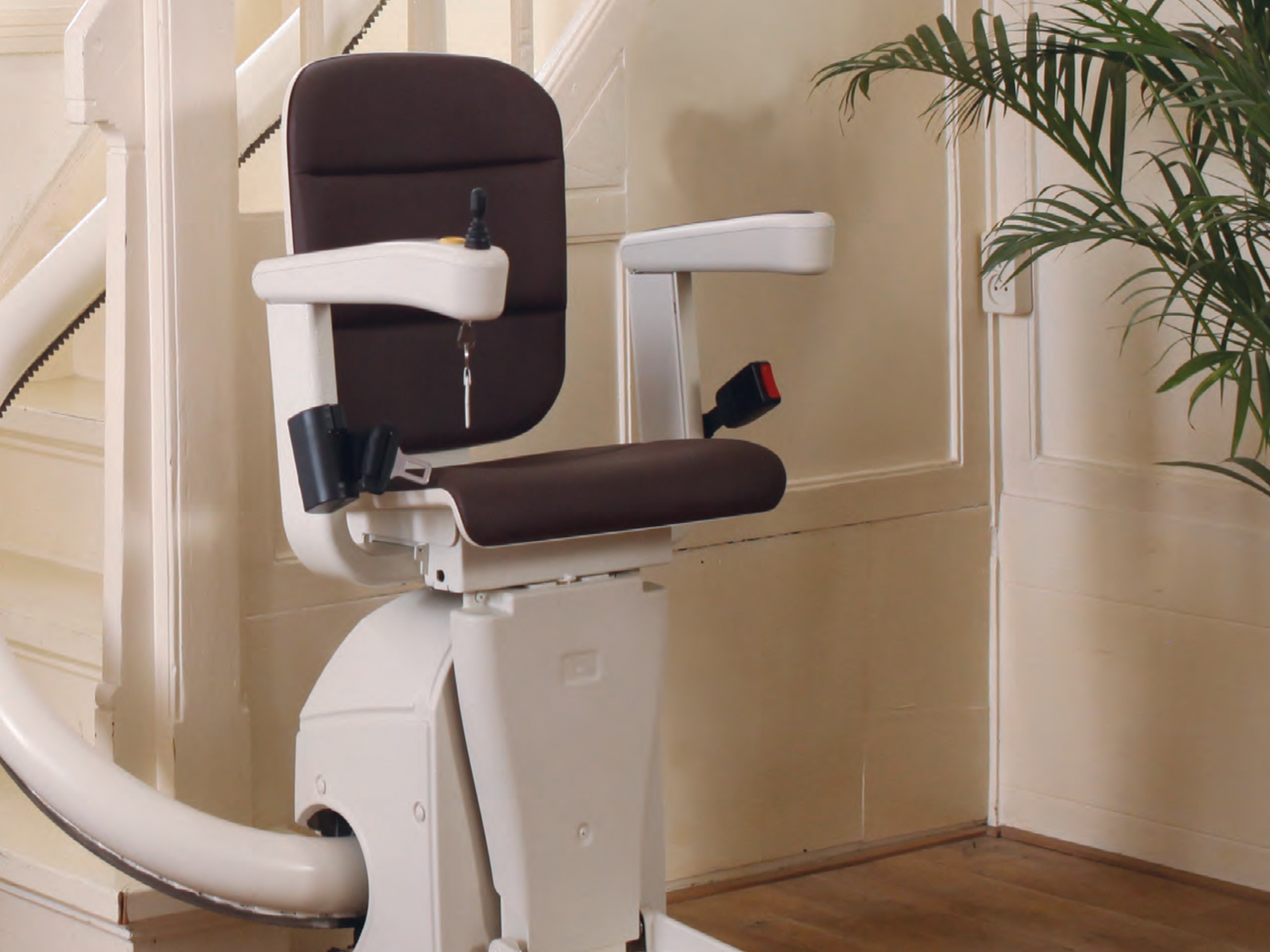 A brown and white stairlift mounted on a curved rail in a home with a wooden floor and a green potted plant nearby.