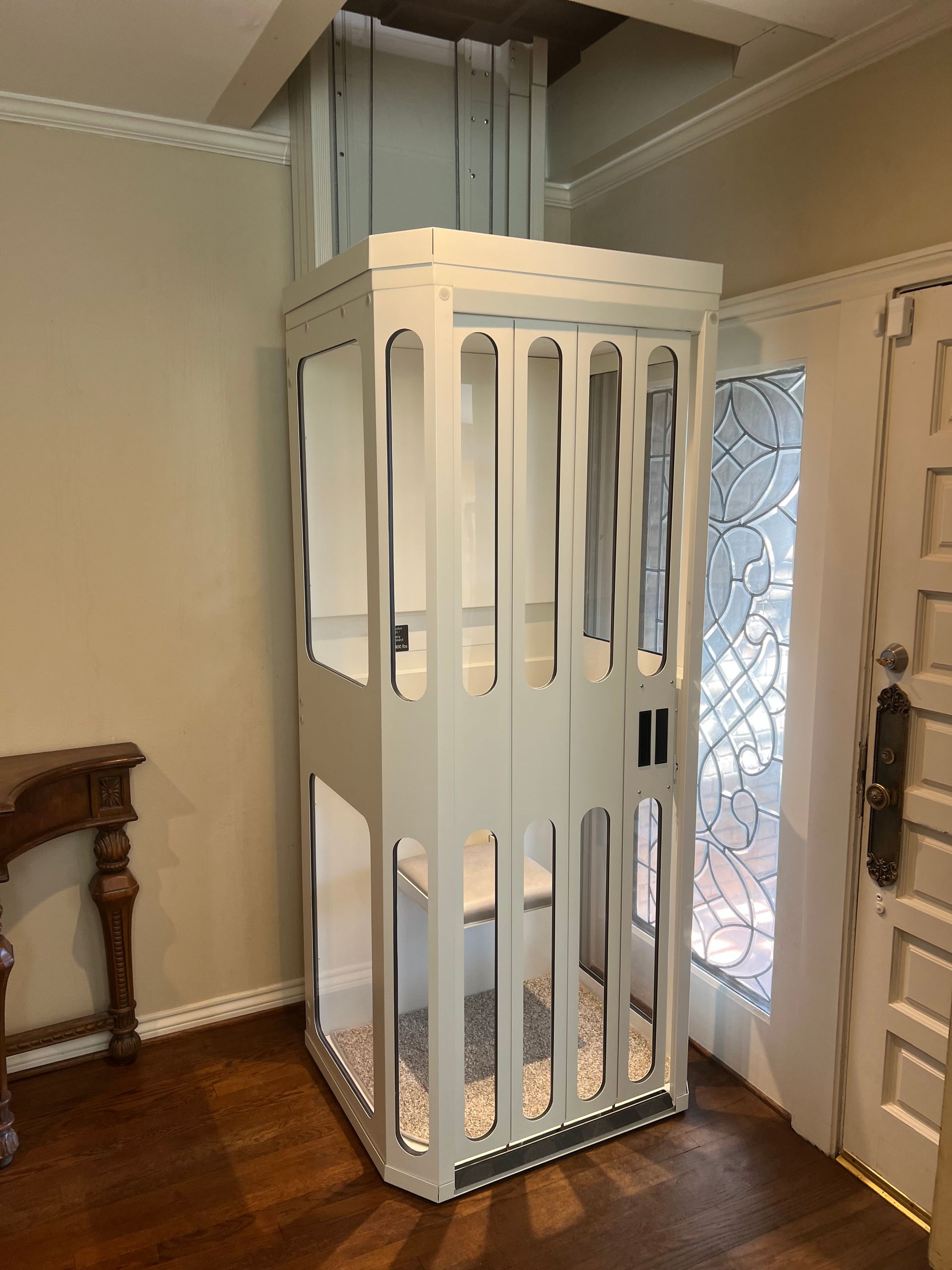 An open home elevator with white paneled walls, wooden flooring, recessed lighting, and a metal control panel.