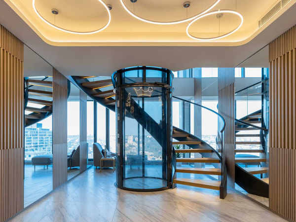 A modern interior featuring a circular glass elevator at the center of a sweeping, wood-trimmed spiral staircase.
