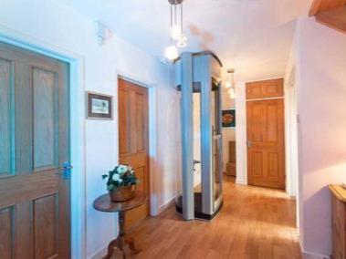 A residential hallway with wooden doors and floors, featuring a modern, cylindrical glass home elevator as a central focus.