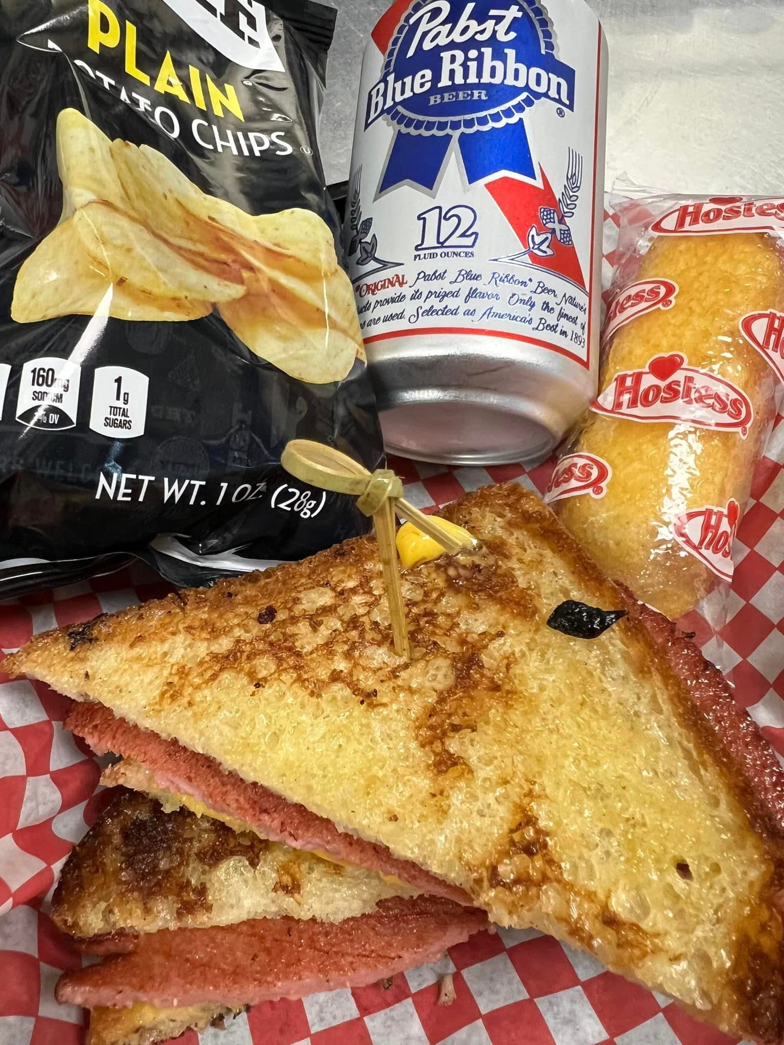 A sandwich , chips , and a can of pabst blue ribbon beer on a checkered table cloth.
