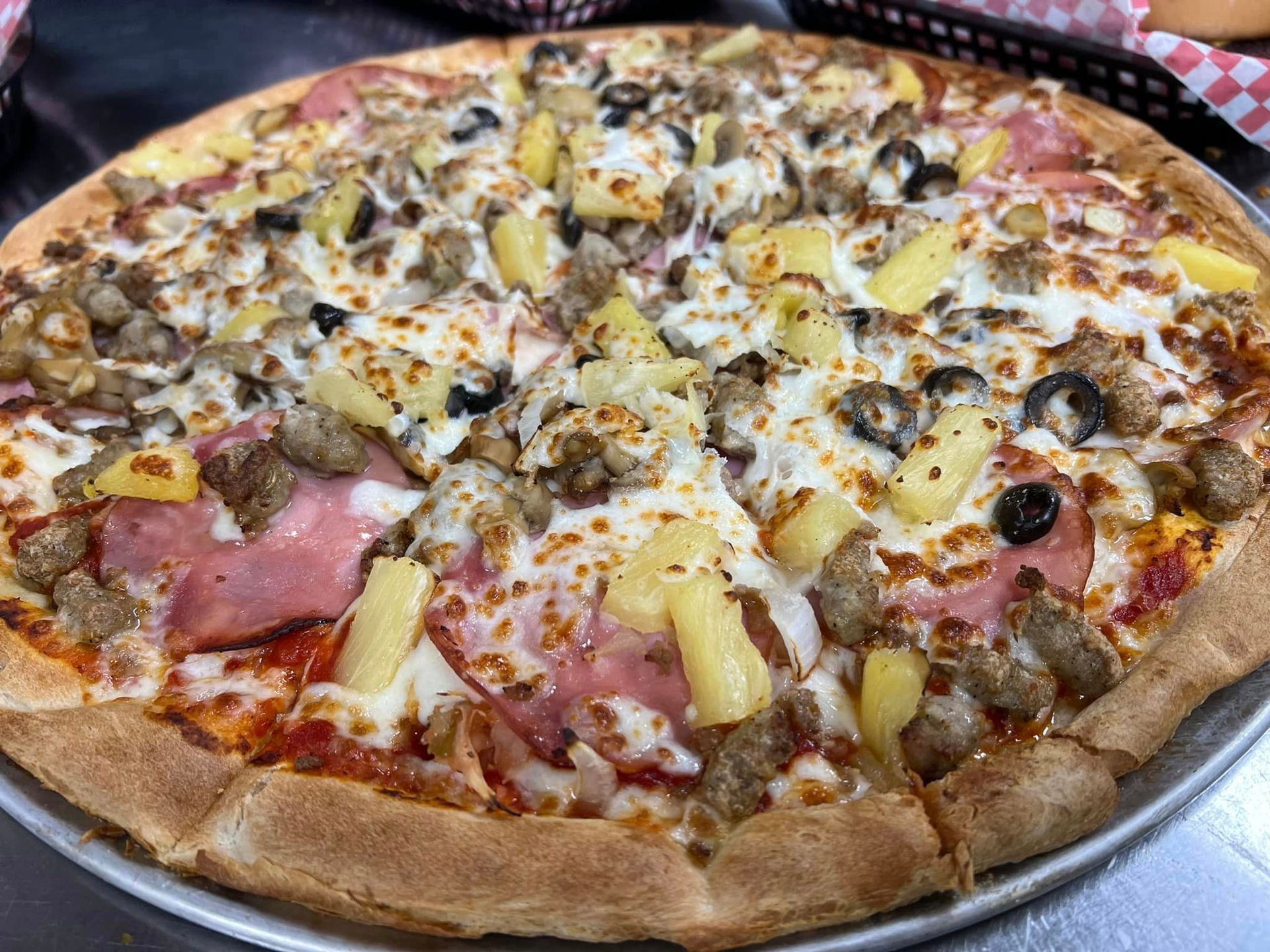 A pizza with pineapple , sausage , olives and ham on a pan on a table.