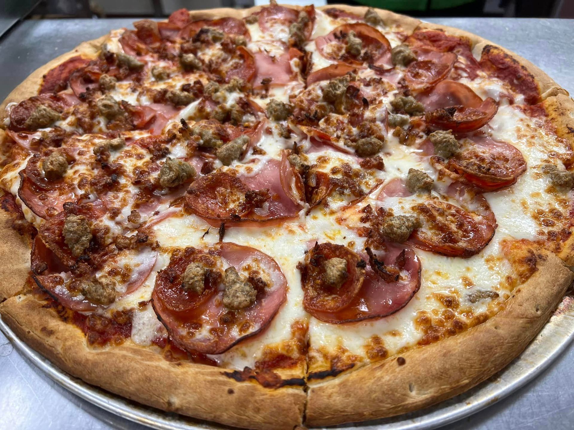 A pizza with ham , sausage , tomatoes and cheese on a pan on a table.