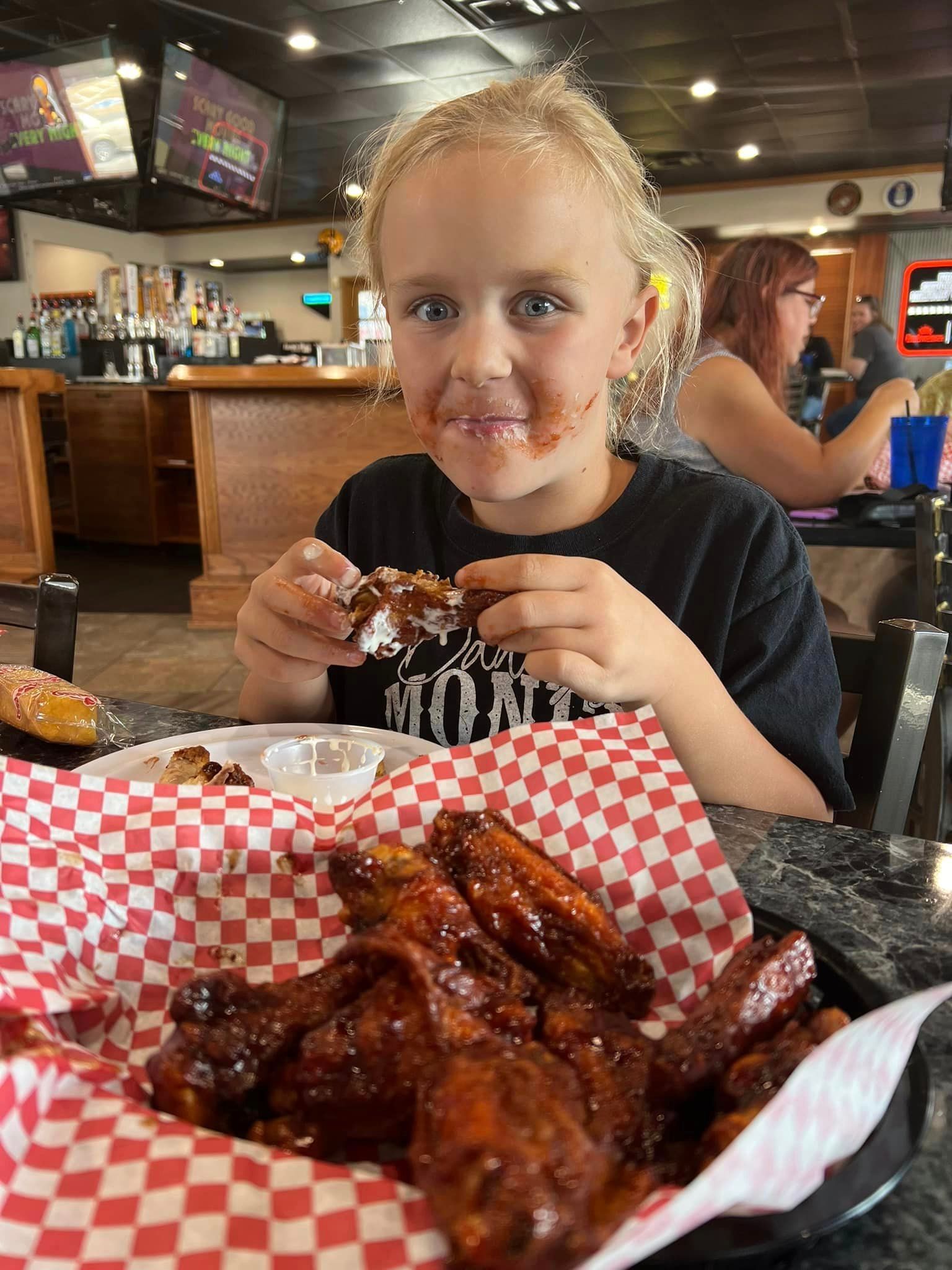 A little girl is sitting at a table eating chicken wings.