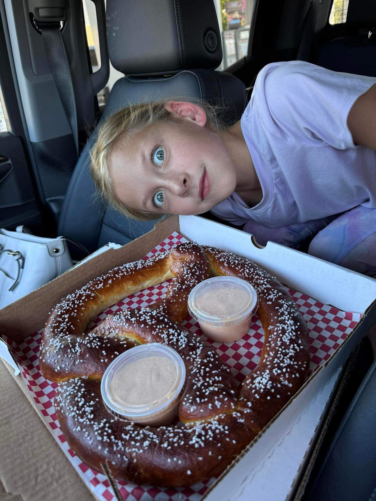 A little girl is sitting in a car next to a box of pretzels.