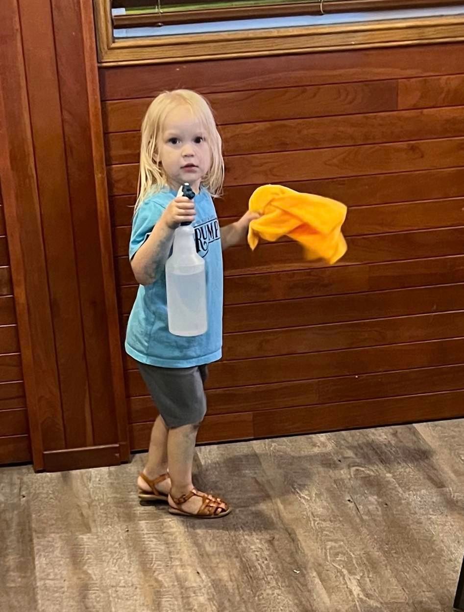 A little girl is standing in a room holding a spray bottle and a towel.