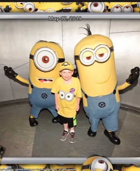 A smiling child in a yellow Minion shirt stands between two Minion costumed characters against a gray paneled background.