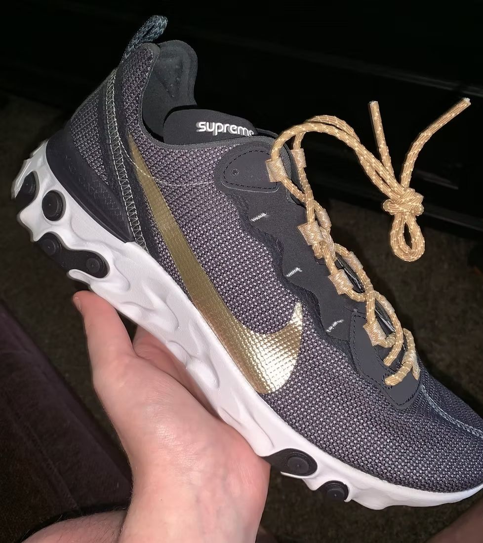A hand holds a gray and gold Nike React Element sneaker with a woven, tan rope-like lace hanging off the side.