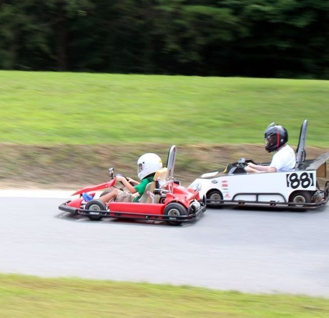 Two people in go-karts racing on an outdoor track; one kart is red and the other is white with the number 88 on its side.