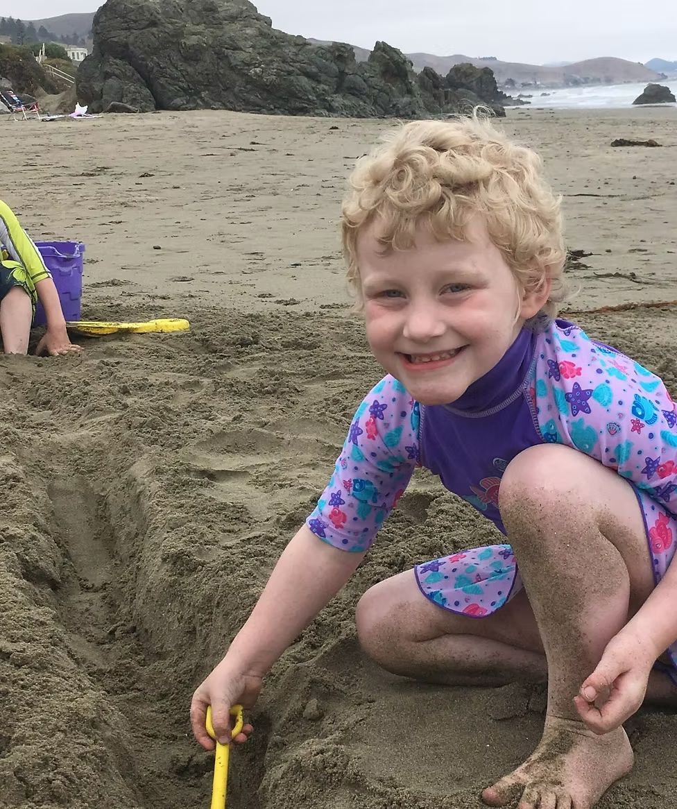 A person with curly blonde hair smiles while digging in the sand at the beach, wearing a colorful, patterned swimsuit.