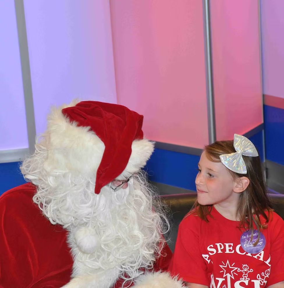 A child wearing a red shirt with a silver bow in their hair looking up at Santa Claus in a festive setting.