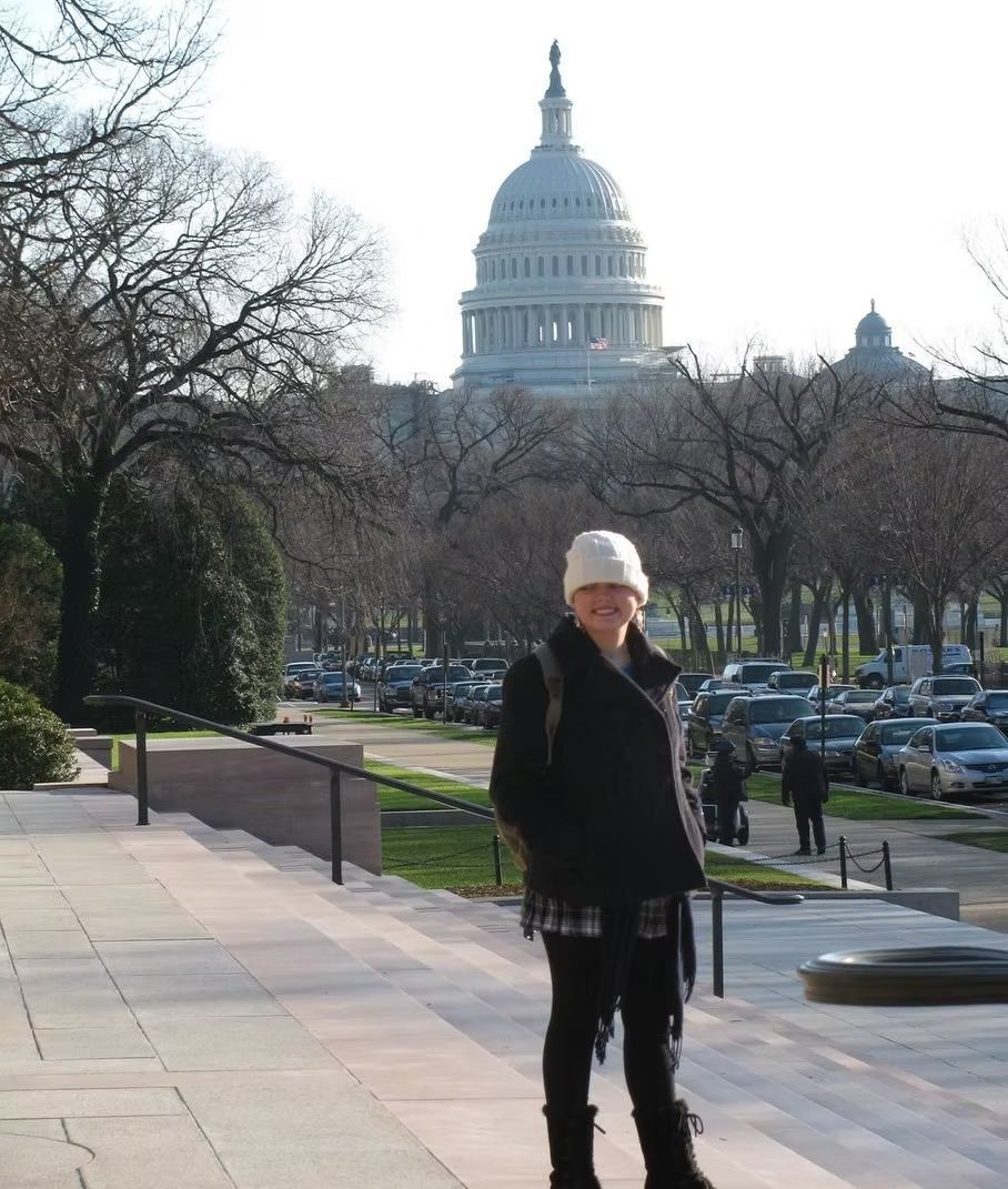A person in a white hat and black coat stands on stone steps with the U.S. Capitol building in the background.