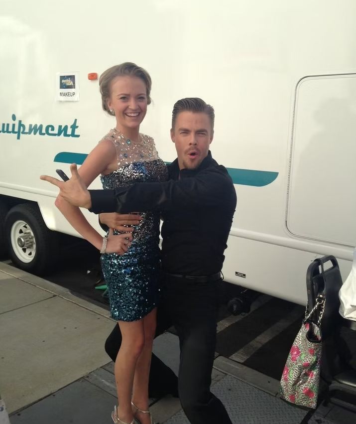 A smiling person in a sparkling blue dress stands next to someone in black making a playful expression by a white truck.