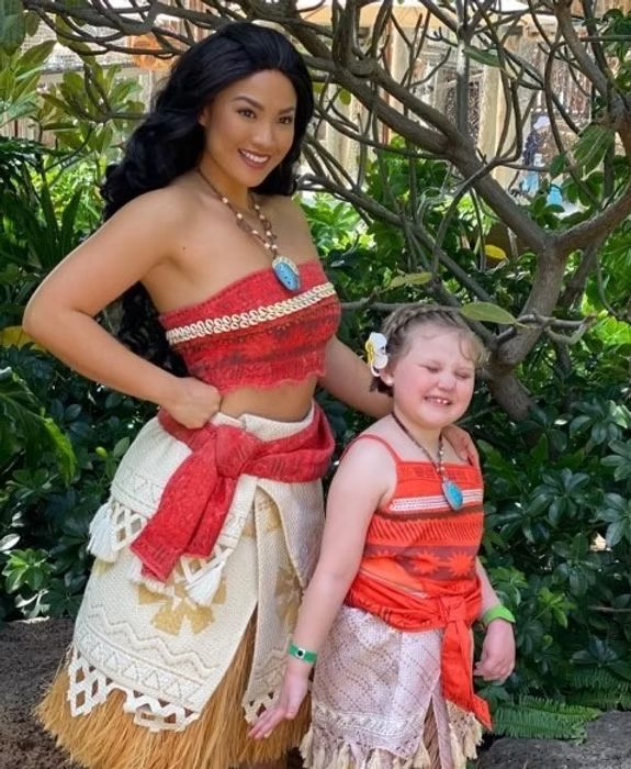 A person dressed as Moana poses with a smiling child wearing a matching costume outdoors among green foliage.