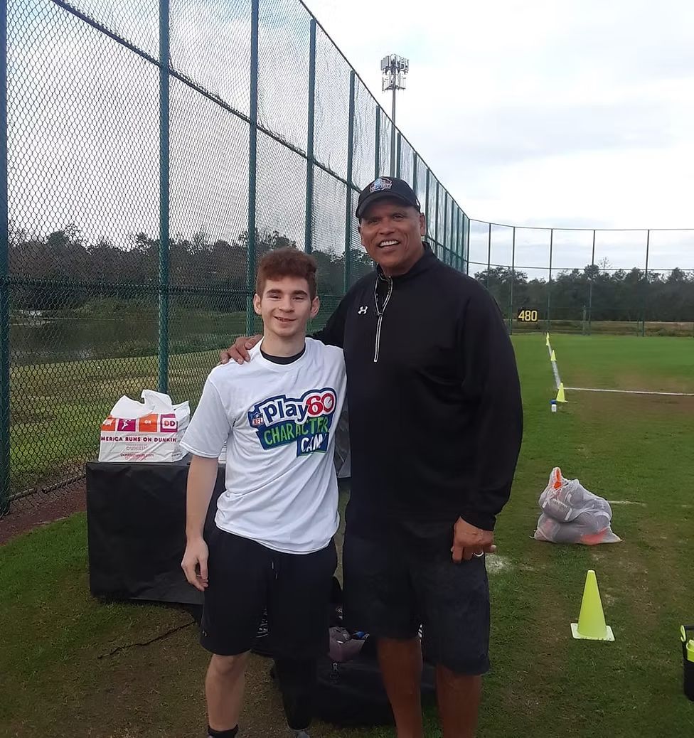 A person in a white t-shirt and an adult in a black jacket stand together on a grassy field by a fence.