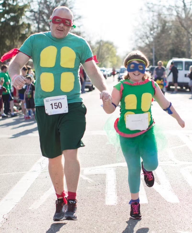 A person and a child dressed as Teenage Mutant Ninja Turtles run side-by-side in a race, wearing masks and race bibs.