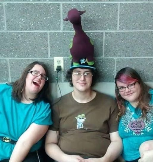 Three friends sit against a grey brick wall; the person in the center wears a stuffed animal balanced on their hat.