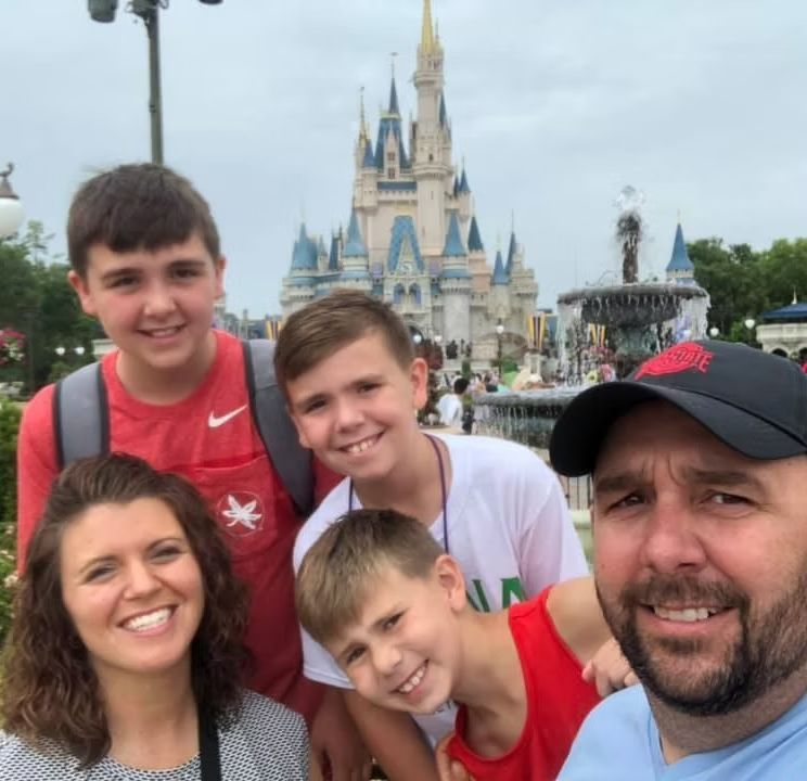 A family poses for a selfie in front of the Cinderella Castle at Disney World, smiling at the camera.