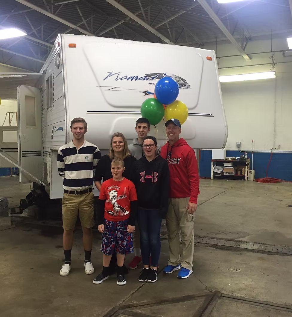 A group of six people stand smiling in front of a white camper trailer indoors, holding a cluster of colorful balloons.