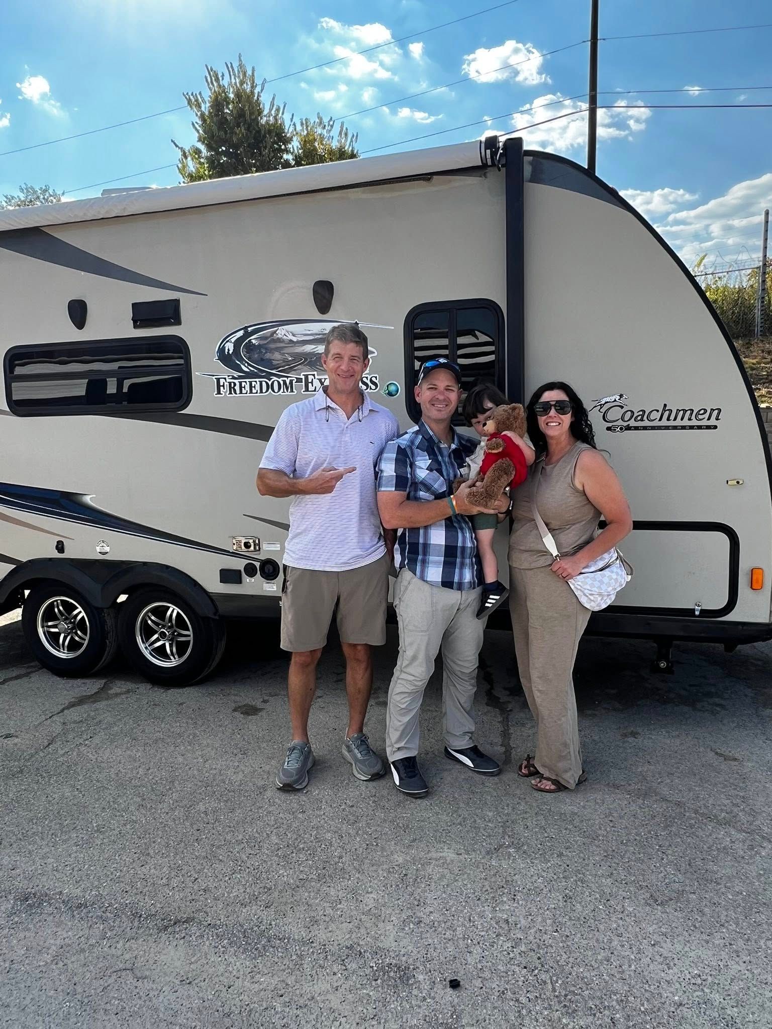 Three people and a small dog stand smiling in front of a tan travel trailer on a paved lot under a blue, sunny sky.