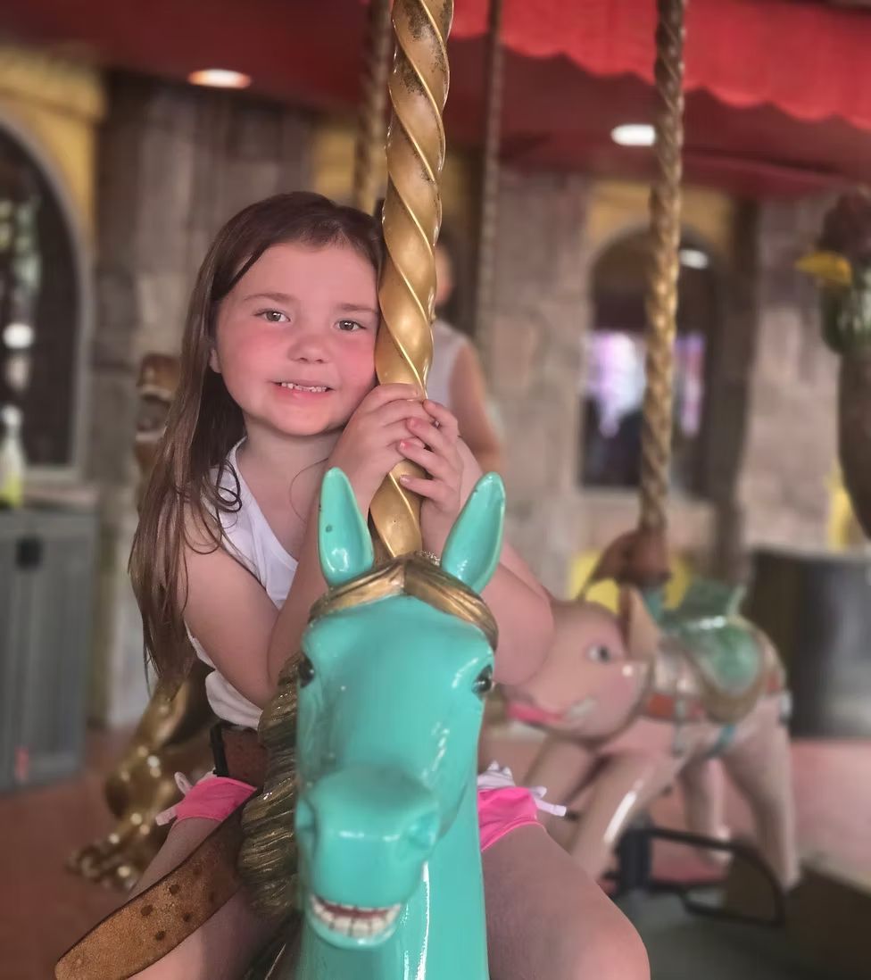 A young child smiles while riding a turquoise carousel horse, holding onto the gold pole.