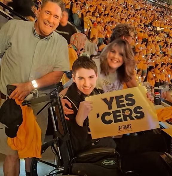 Three people smile in a crowded arena while the person in front holds a yellow sign that reads, 