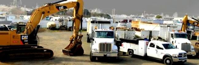 A yellow Caterpillar excavator is operating near various white trucks and equipment in a construction site yard.