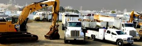 A yellow Caterpillar excavator is operating near various white trucks and equipment in a construction site yard.