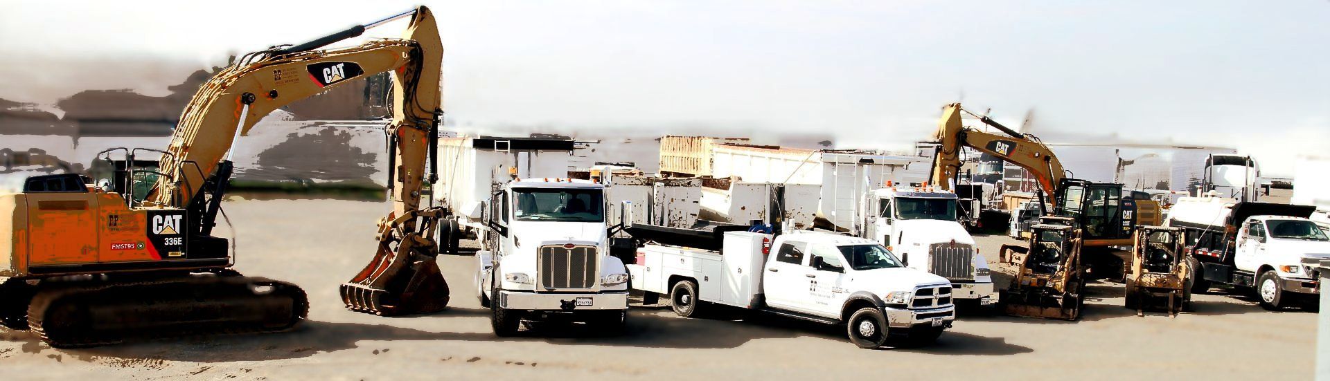 Construction vehicles parked on a sandy, overcast day. Includes excavators, trucks, and other heavy equipment.