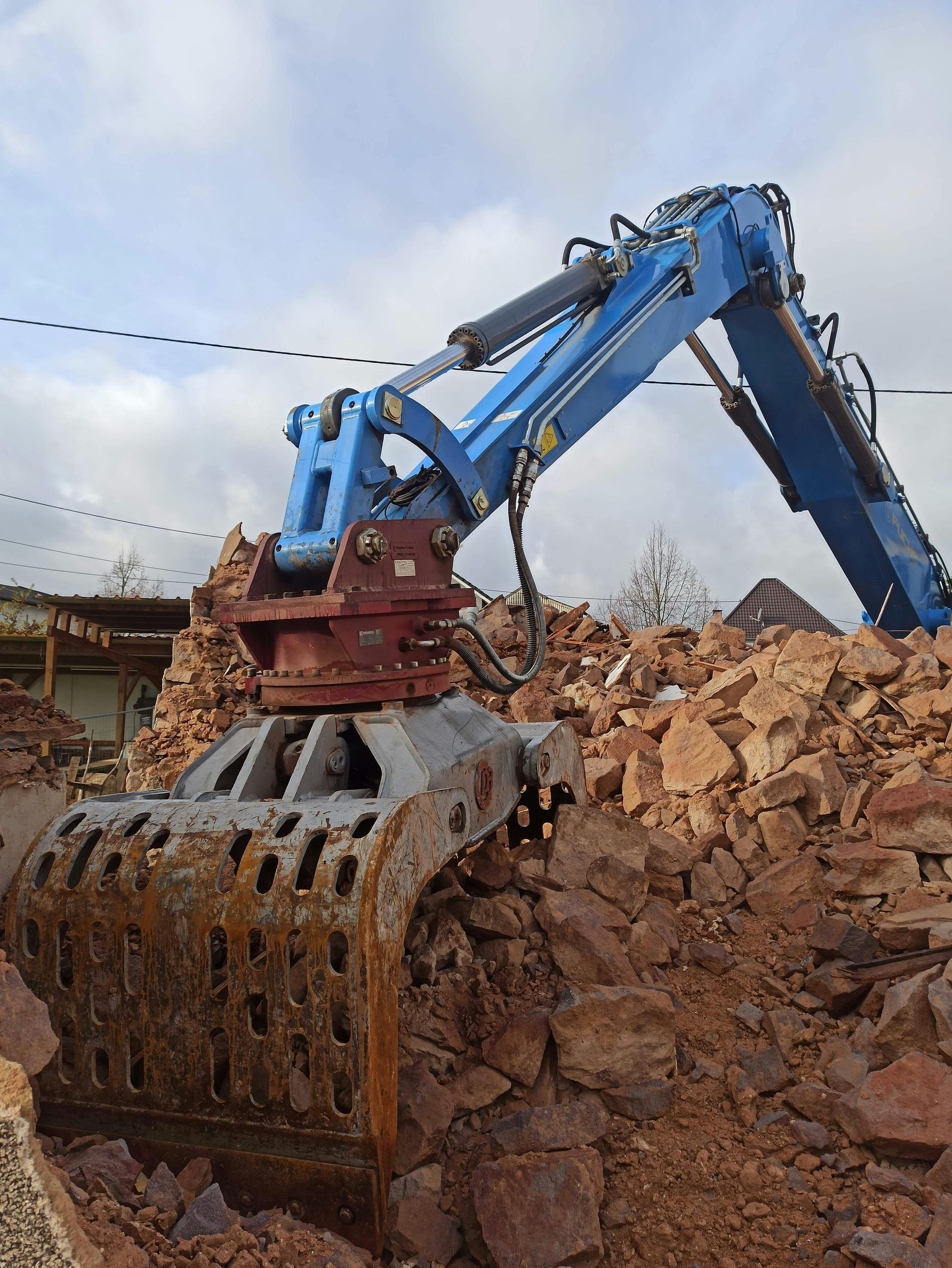 Blue excavator with a demolition attachment demolishing a brick structure against a cloudy sky.