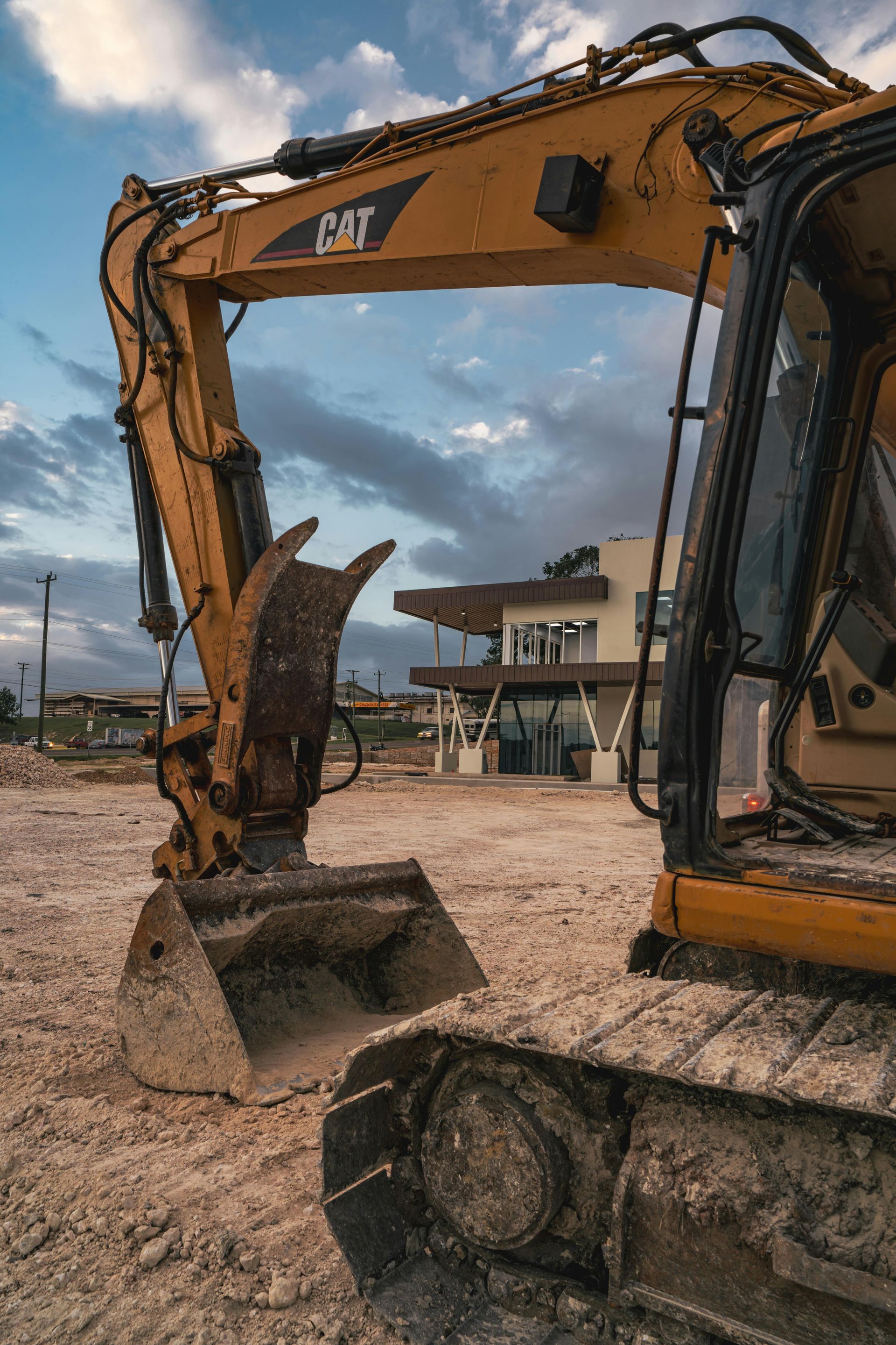 Yellow Caterpillar excavator at a construction site. The machine's arm extends towards a building under a cloudy sky.