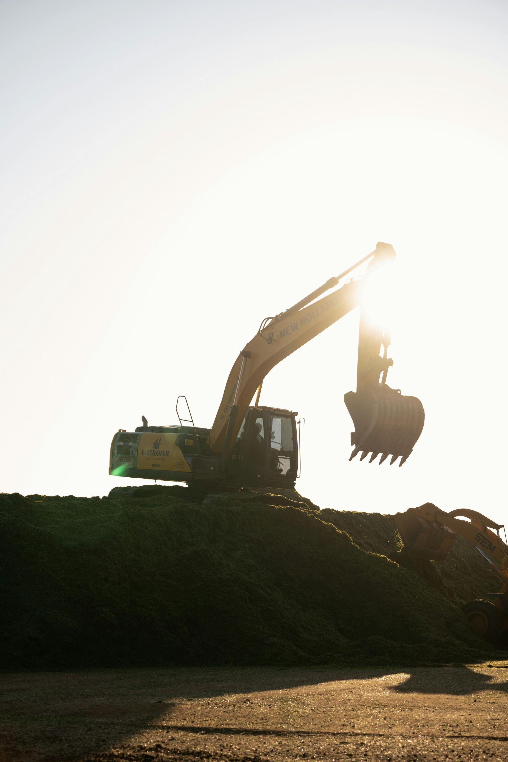 An excavator silhouetted against the sun, working on a pile of dark material, possibly mulch or soil.