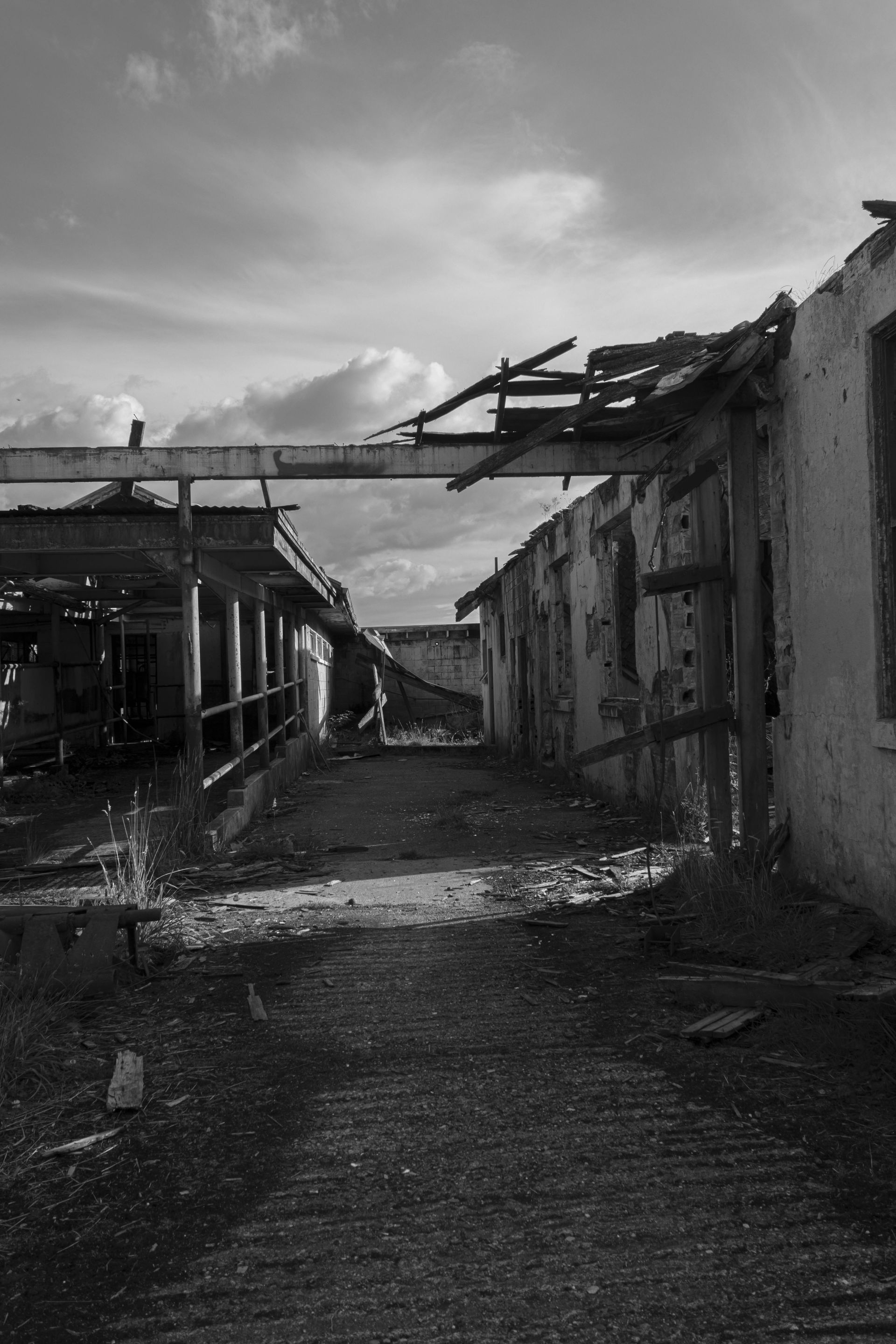 Black and white photo of a dilapidated building with a path leading through it. The walls are damaged, and the sky is cloudy.