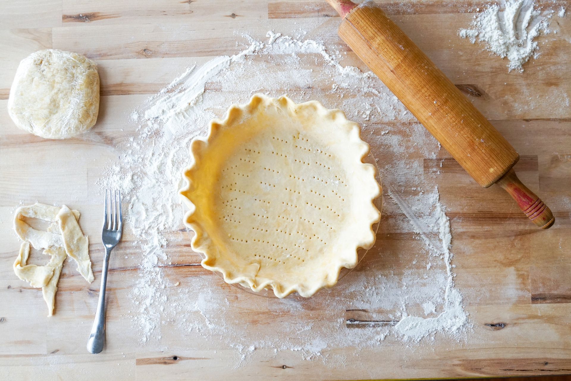 A pie crust is sitting on a wooden table next to a rolling pin and a fork.